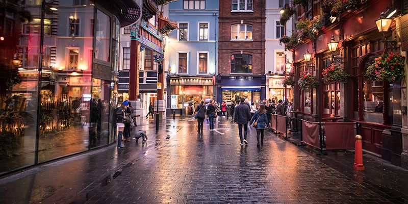 People walking down a wet, lit city street surrounded by shops and restaurants at dusk.