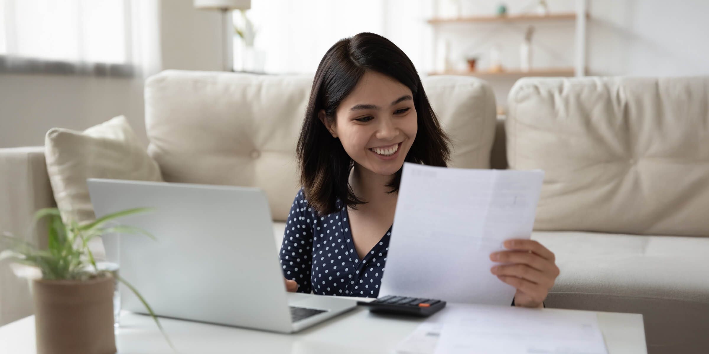 woman smiling working at laptop and looking at piece of paper in her hand