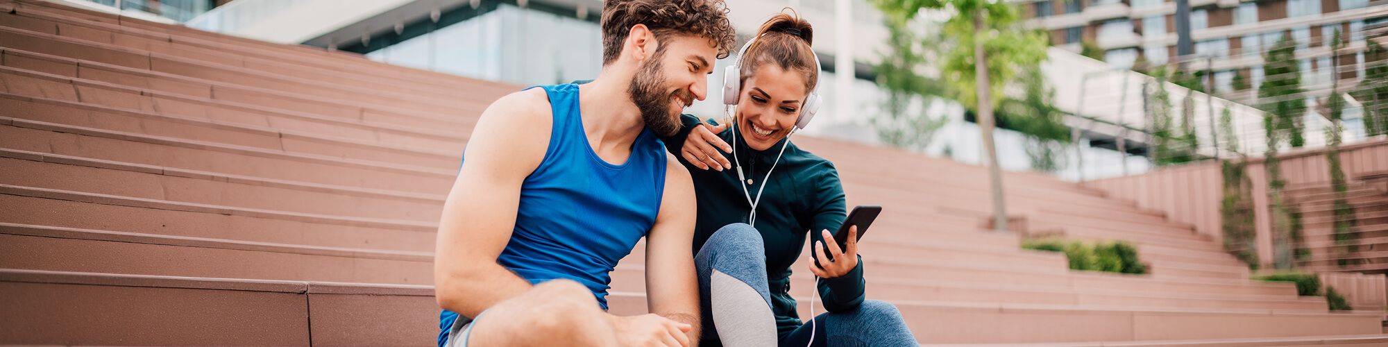 man and woman in athletic wear sitting in empty stadium bleachers and smiling while looking at phone