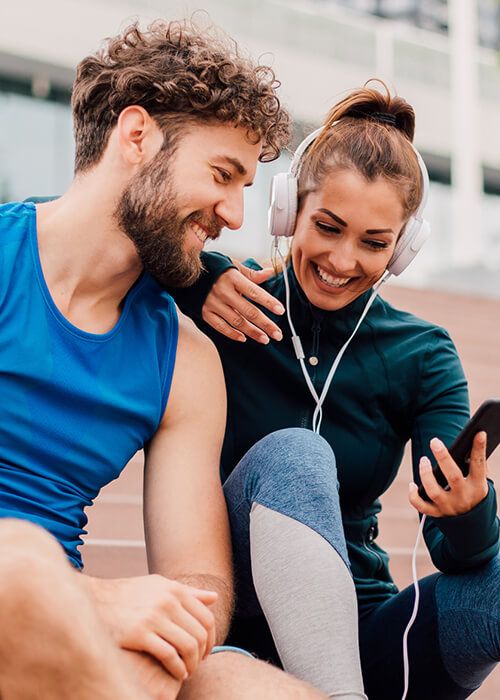 man and woman in athletic wear sitting in empty stadium bleachers and smiling while looking at phone