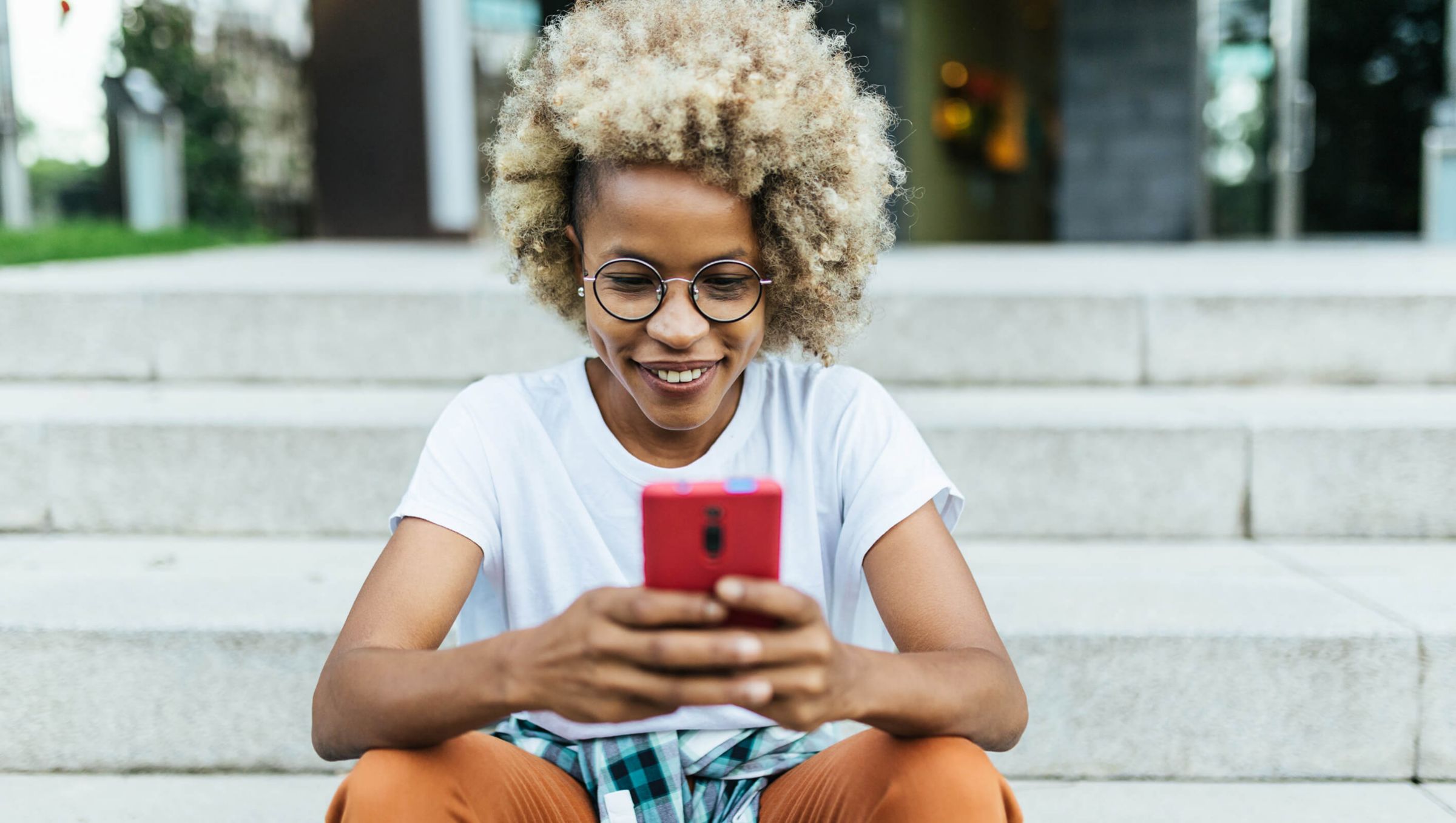 A person with curly blonde hair sits on outdoor stairs, intently looking at a smartphone