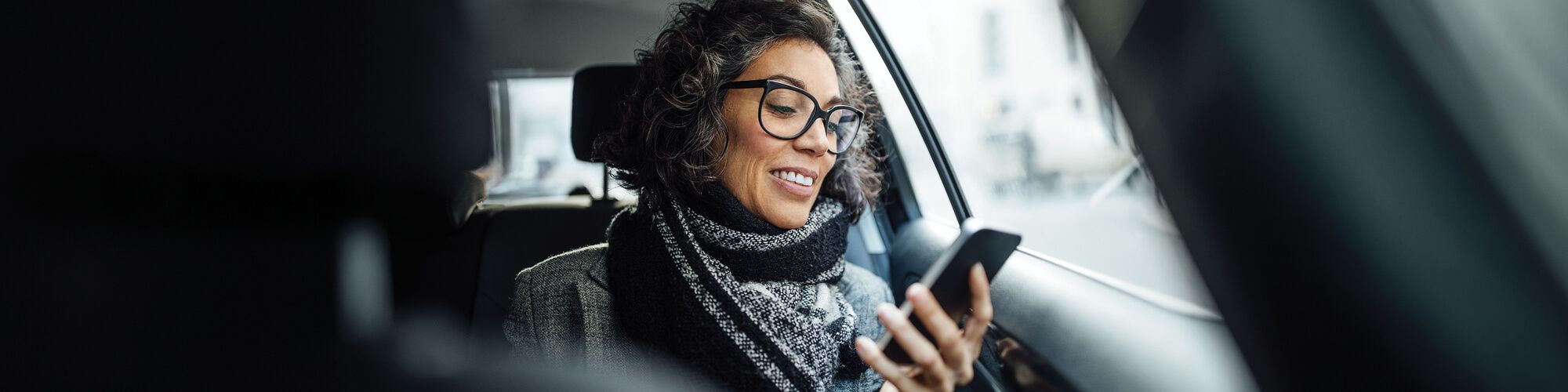 woman wearing a warm scarf and eye glasses sitting in car smiling while looking at phone