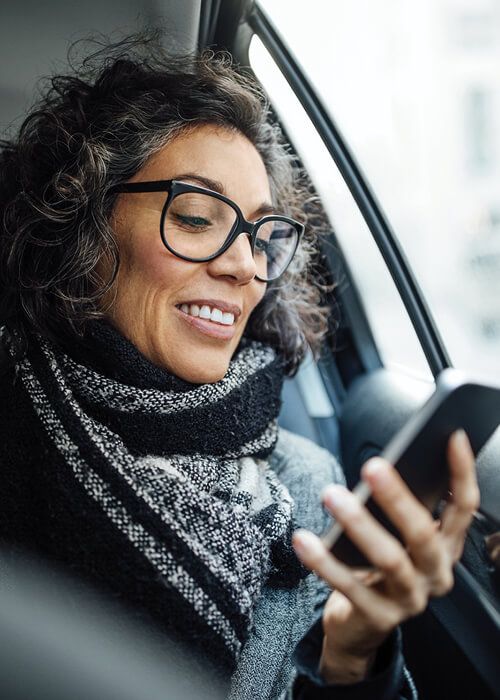 woman wearing a warm scarf and eye glasses sitting in car smiling while looking at phone
