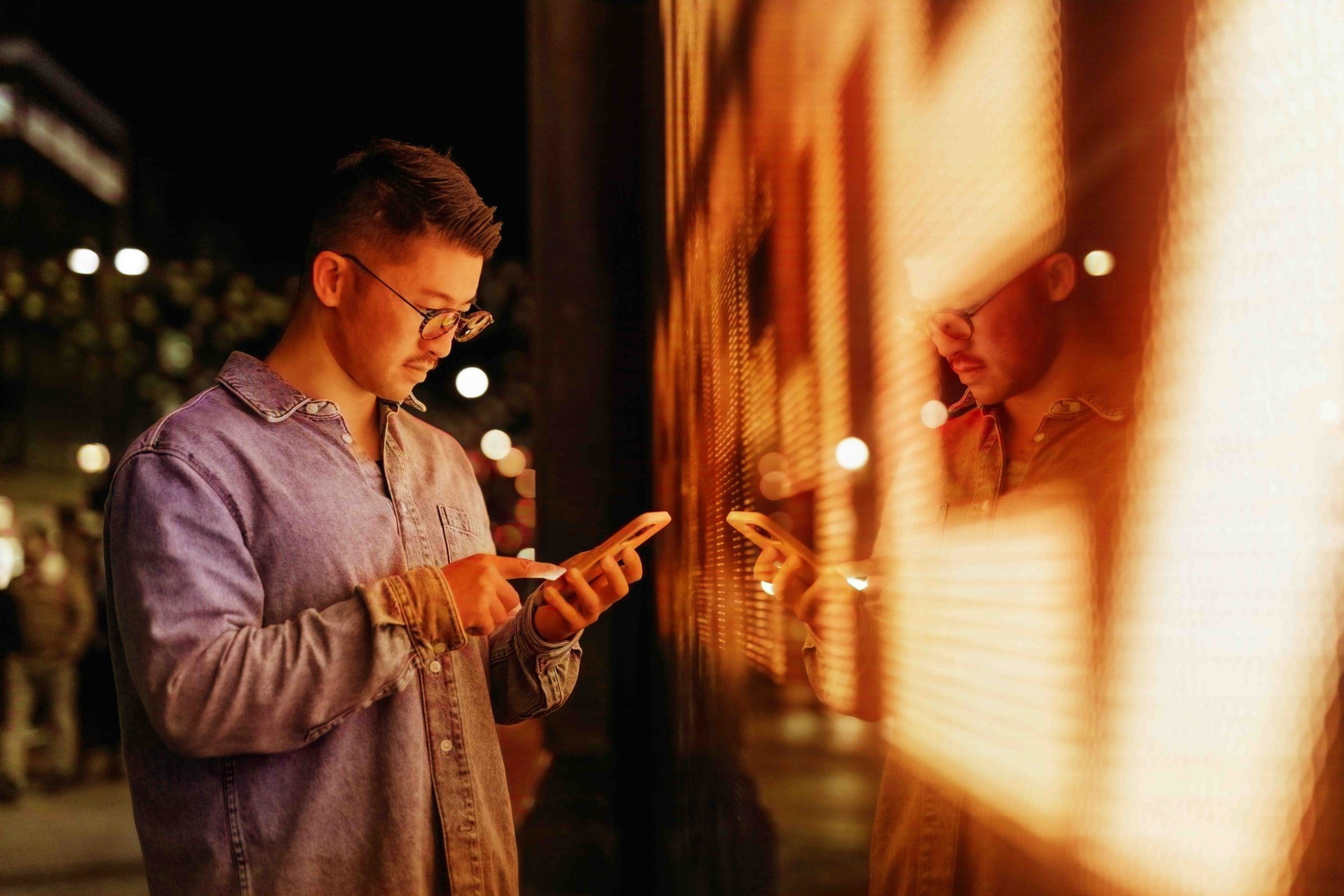 Side view of a young Asian man using online banking app on smartphone, standing next to an illuminated red LED & digital display screens in the city at night. Mobile technology in everyday life.