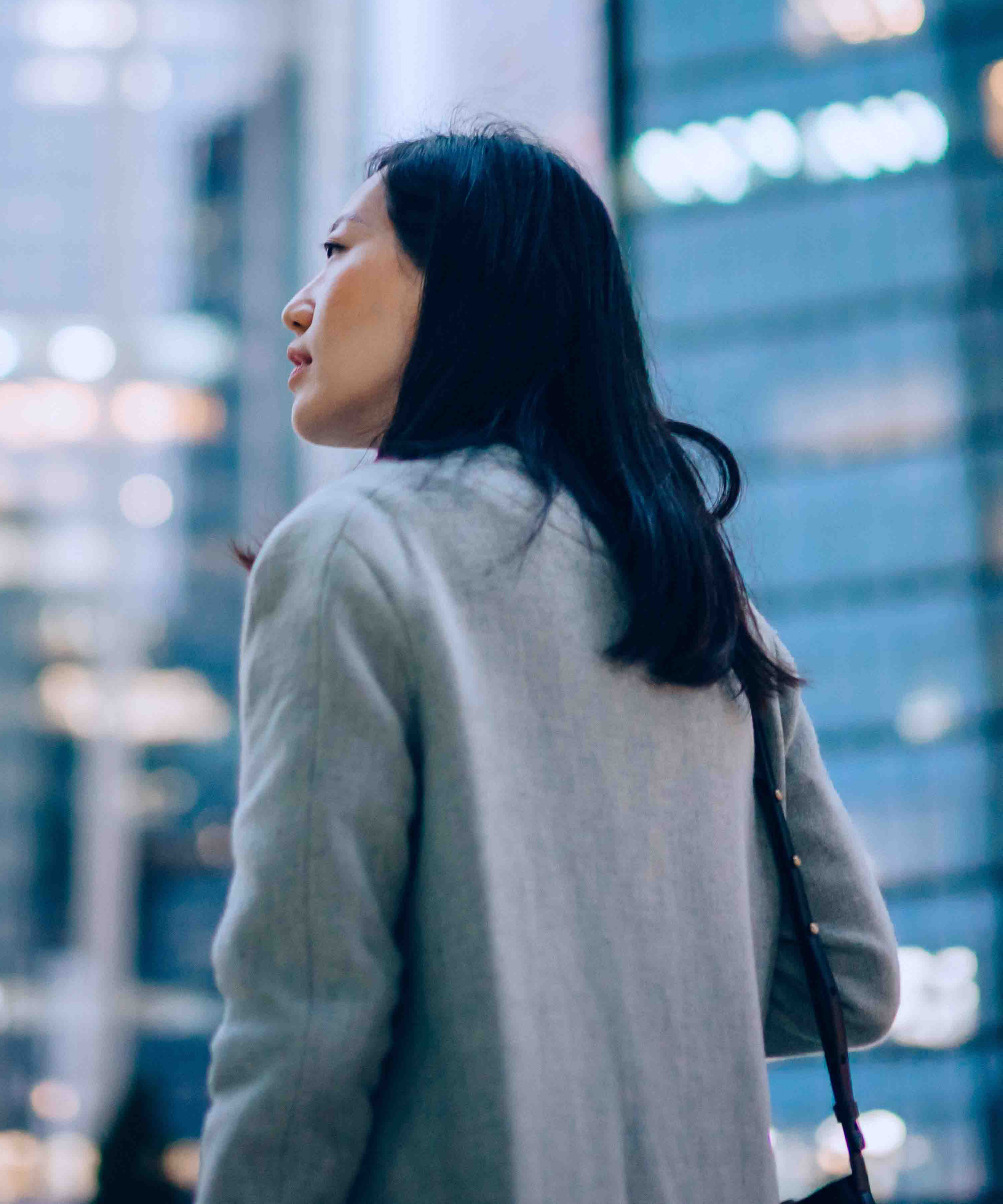 Low angle side profile of confident and professional young Asian businesswoman looking up while standing against contemporary corporate skyscrapers with illuminated facade in financial district in the evening. Female leadership and determined to success.