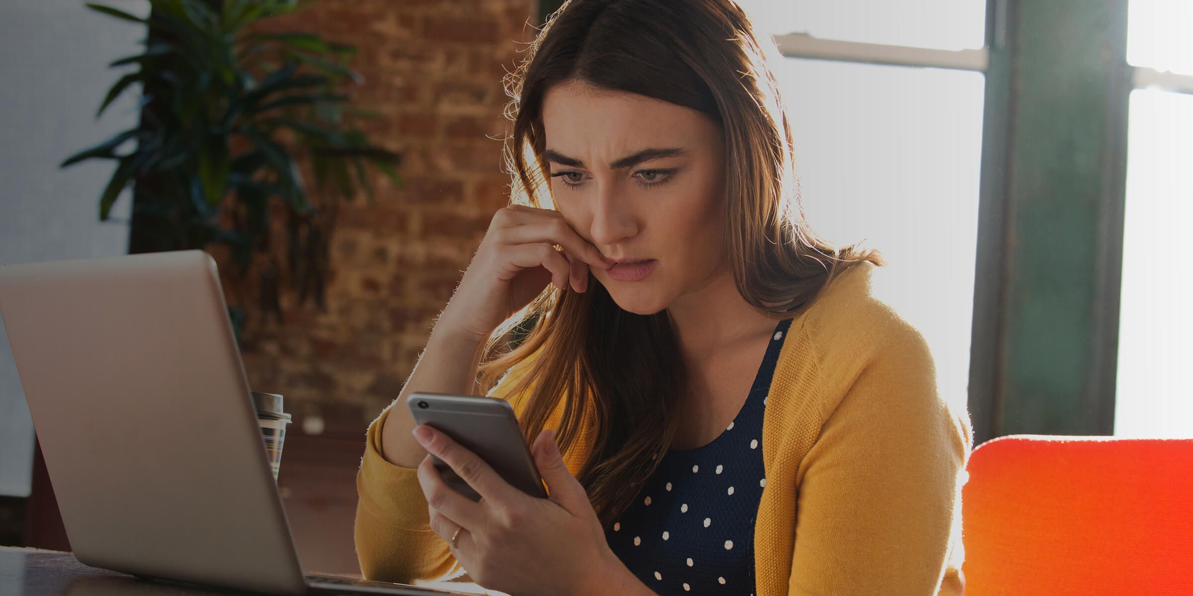 Woman in a yellow cardigan using smartphone with laptop on desk, resting on a purple blanket