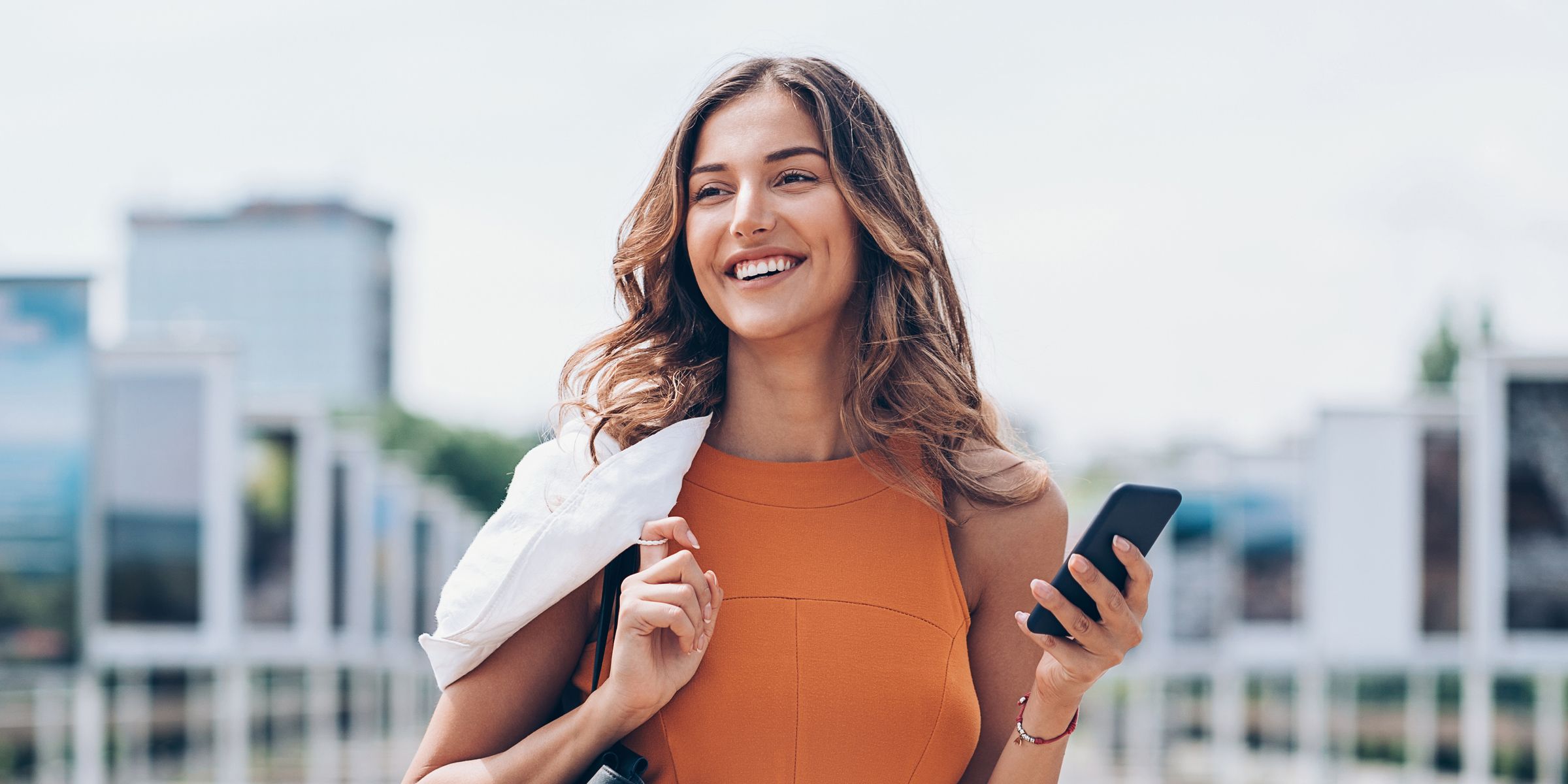Smiling woman in an orange top holding a phone while walking outdoors.