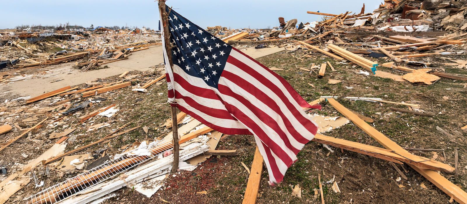  A dramatic, low-angle photograph shows an American flag planted on a makeshift wooden pole amid a massive field of wreckage. The ground is covered in splintered wood, debris, and rubble from destroyed buildings, suggesting the aftermath of a severe natural disaster, likely a tornado or hurricane. The flag is rippling in the wind, a symbol of resilience contrasting with the widespread devastation in the background.