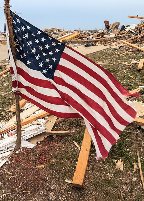  A dramatic, low-angle photograph shows an American flag planted on a makeshift wooden pole amid a massive field of wreckage. The ground is covered in splintered wood, debris, and rubble from destroyed buildings, suggesting the aftermath of a severe natural disaster, likely a tornado or hurricane. The flag is rippling in the wind, a symbol of resilience contrasting with the widespread devastation in the background.