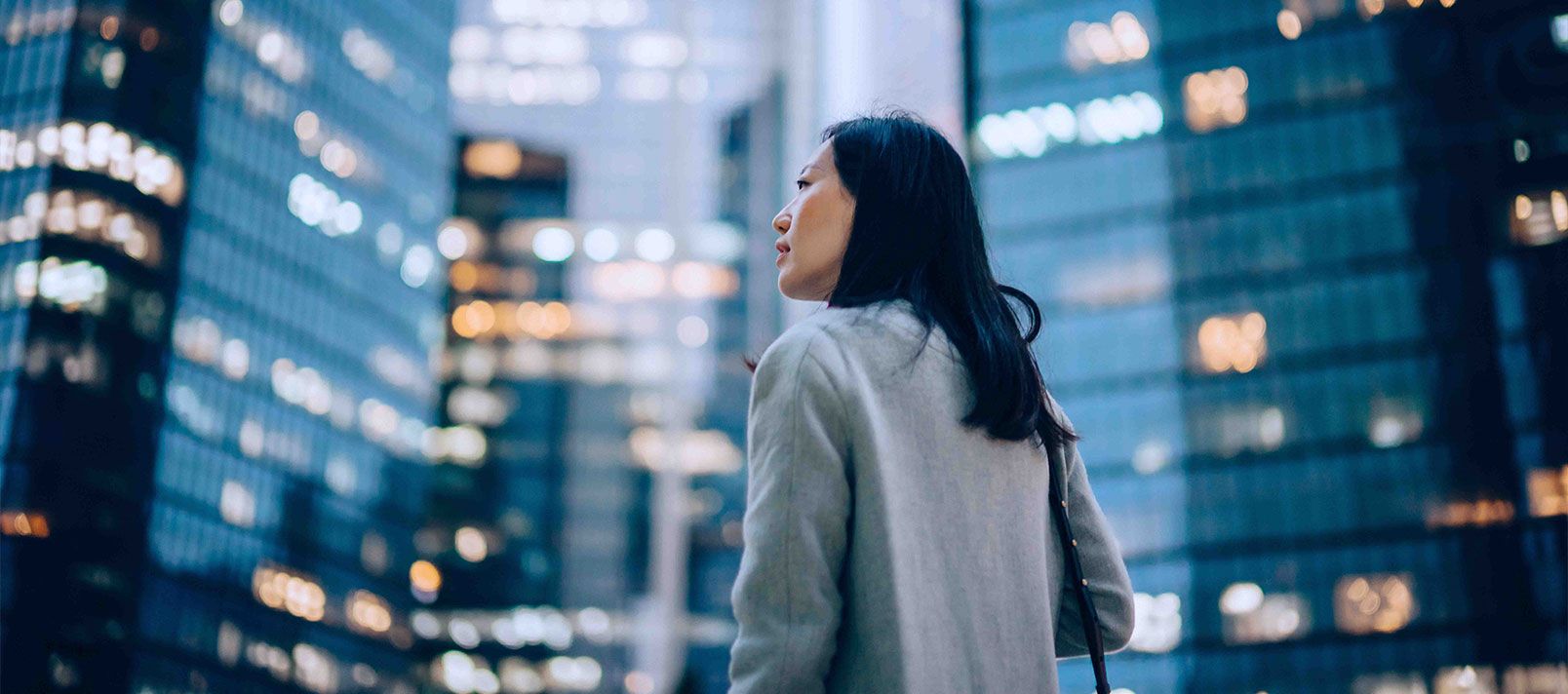 Low angle side profile of confident and professional young Asian businesswoman looking up while standing against contemporary corporate skyscrapers with illuminated facade in financial district in the evening. Female leadership and determined to success.