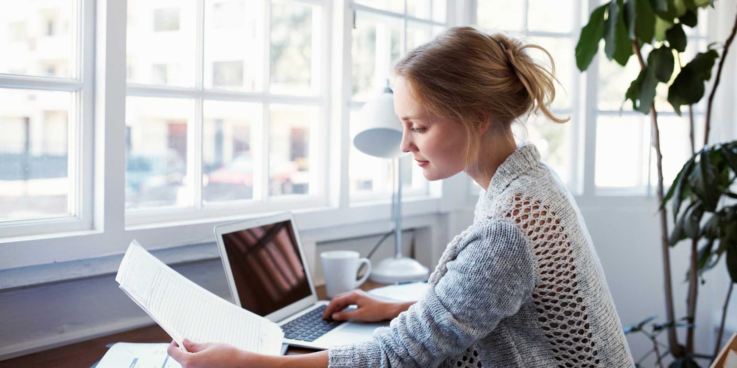 a woman sitting at a laptop desk and looking at a paper